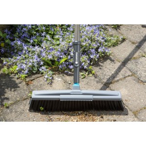 Scopa per esterni Gardena grigia con manico davanti a piante fiorite su pavimentazione.