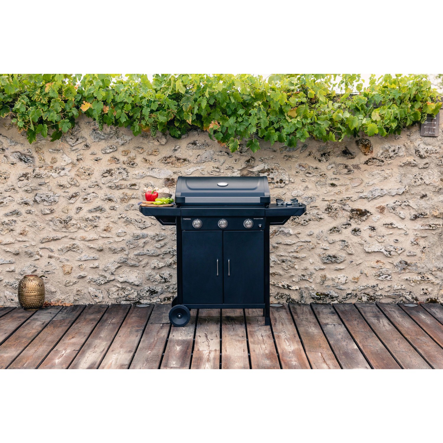 Barbecue a gas nero t.b.t. con ripiani e mobile base davanti a muro di pietra.