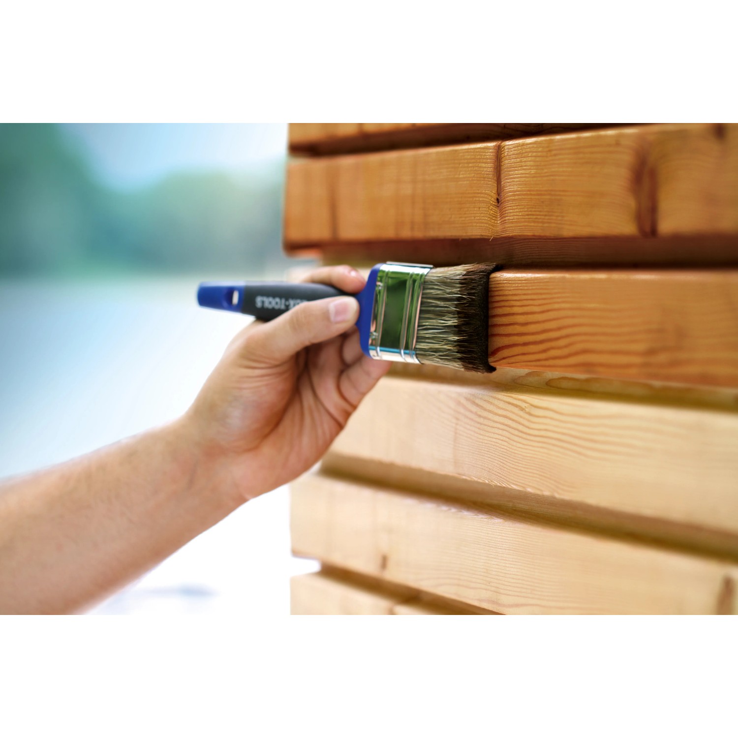 Mano che vernicia il legno con pennello piatto LUX Soft Grip Varnish 70 mm. Pennello con manico blu.