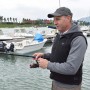 Uomo che pesca con gilet da lavoro t.b.t. e barche sullo sfondo.