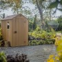 Casetta da giardino in plastica marrone con finestra e porta in un ambiente da giardino.