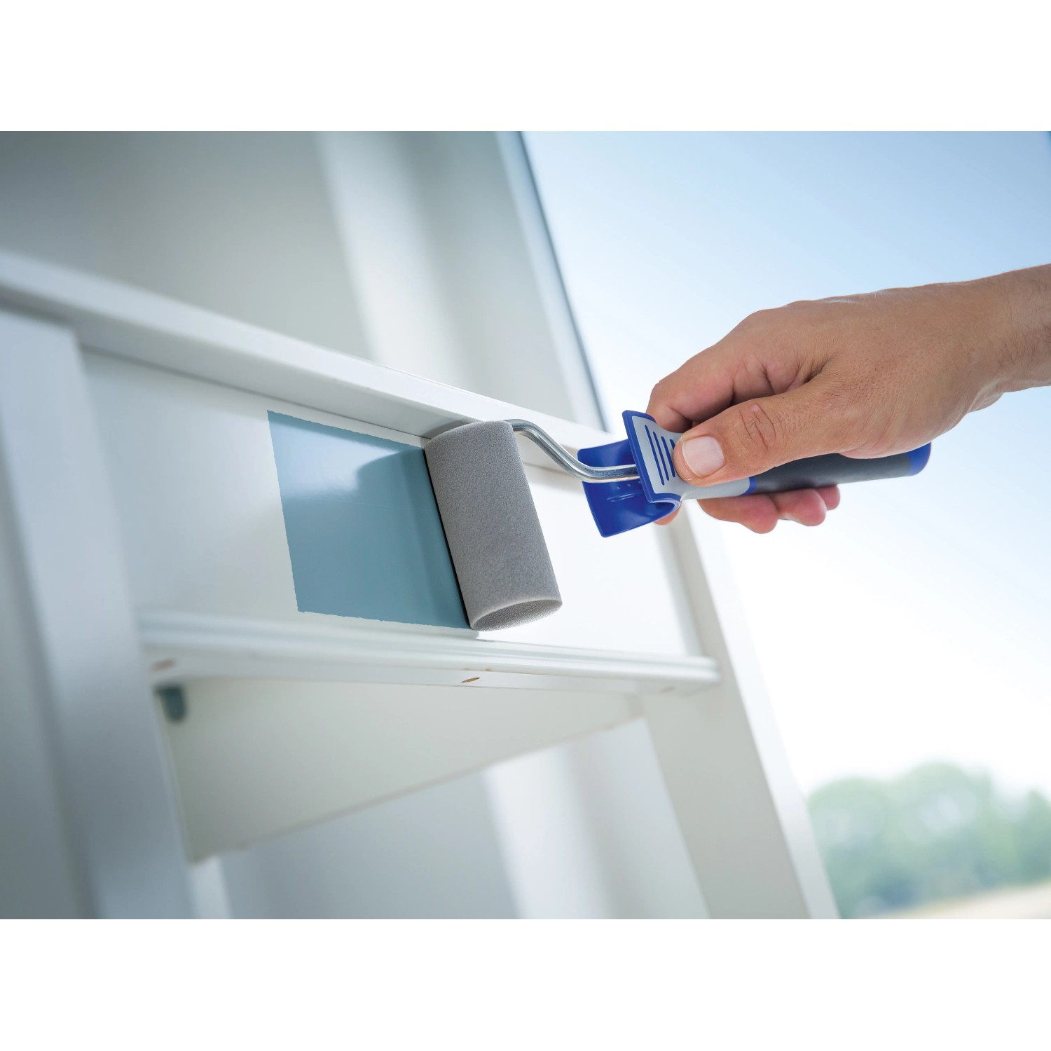 Mano che dipinge di blu un davanzale bianco con rullo in schiuma LUX, 7 cm, concavo. Set di rulli per pittura, 2 pezzi.