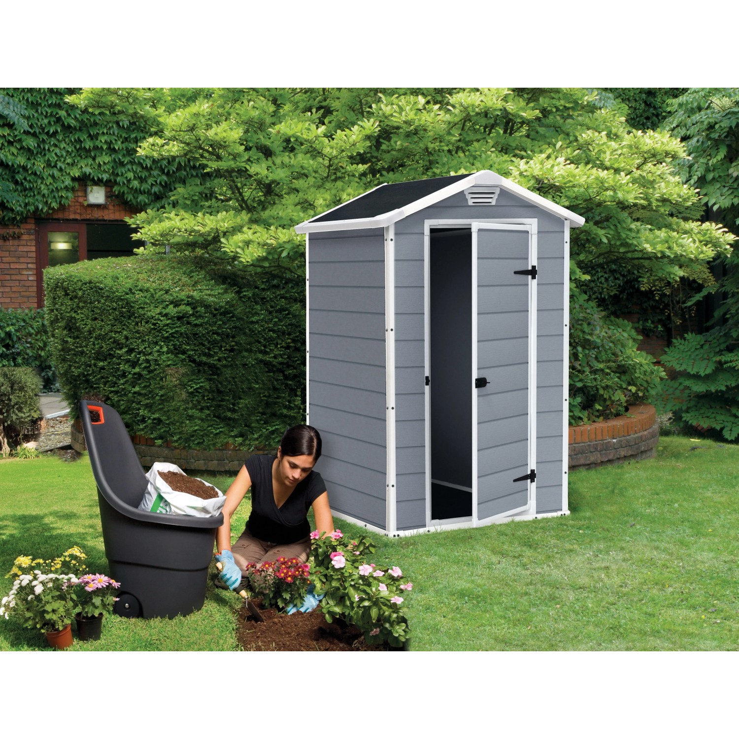 Casetta da giardino Keter Manor 4x3 grigio-bianca con porta aperta e donna che pianta un'aiuola.