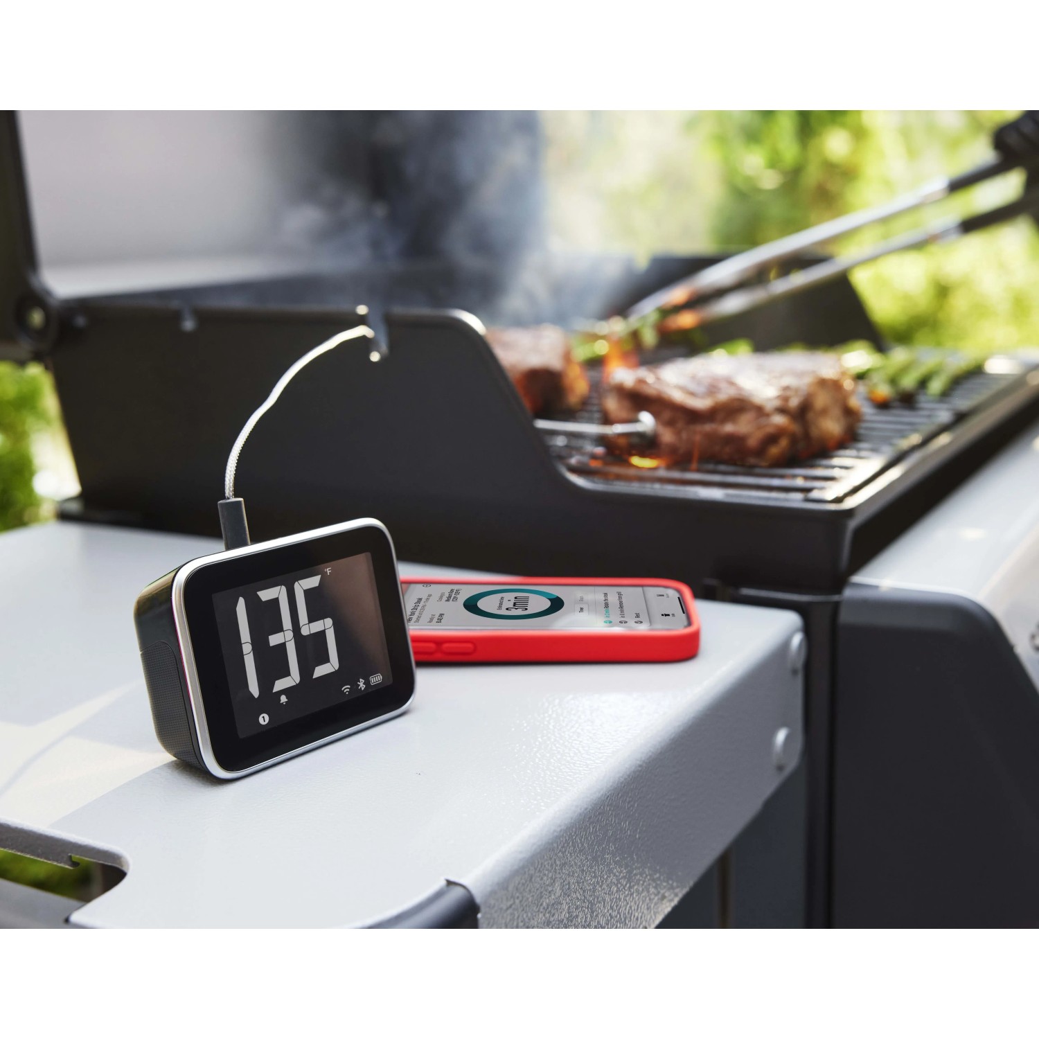 Base display Weber Smart Hub nero-argento con sensore temperatura e smartphone vicino al barbecue.