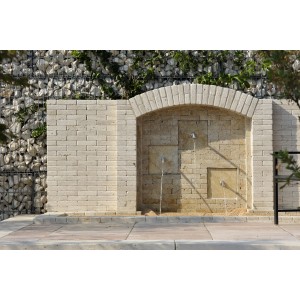 Fontana in pietra con tre bocche d'acqua davanti a muro di gabbioni.
