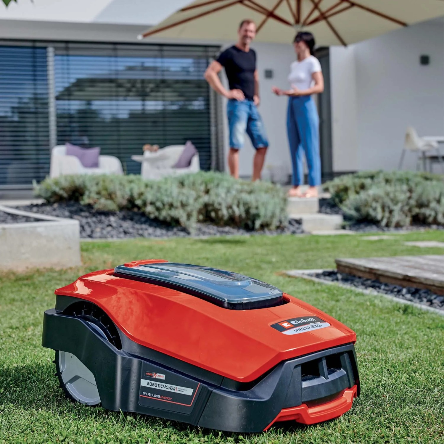 Robot tagliaerba t.b.t. rosso su prato davanti a casa con persone sullo sfondo.