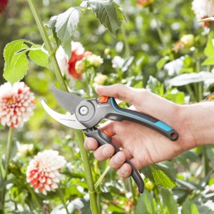 Mano che tiene le cesoie da giardino Gardena B/M davanti a un giardino fiorito. Cesoie bypass con manico blu-grigio.