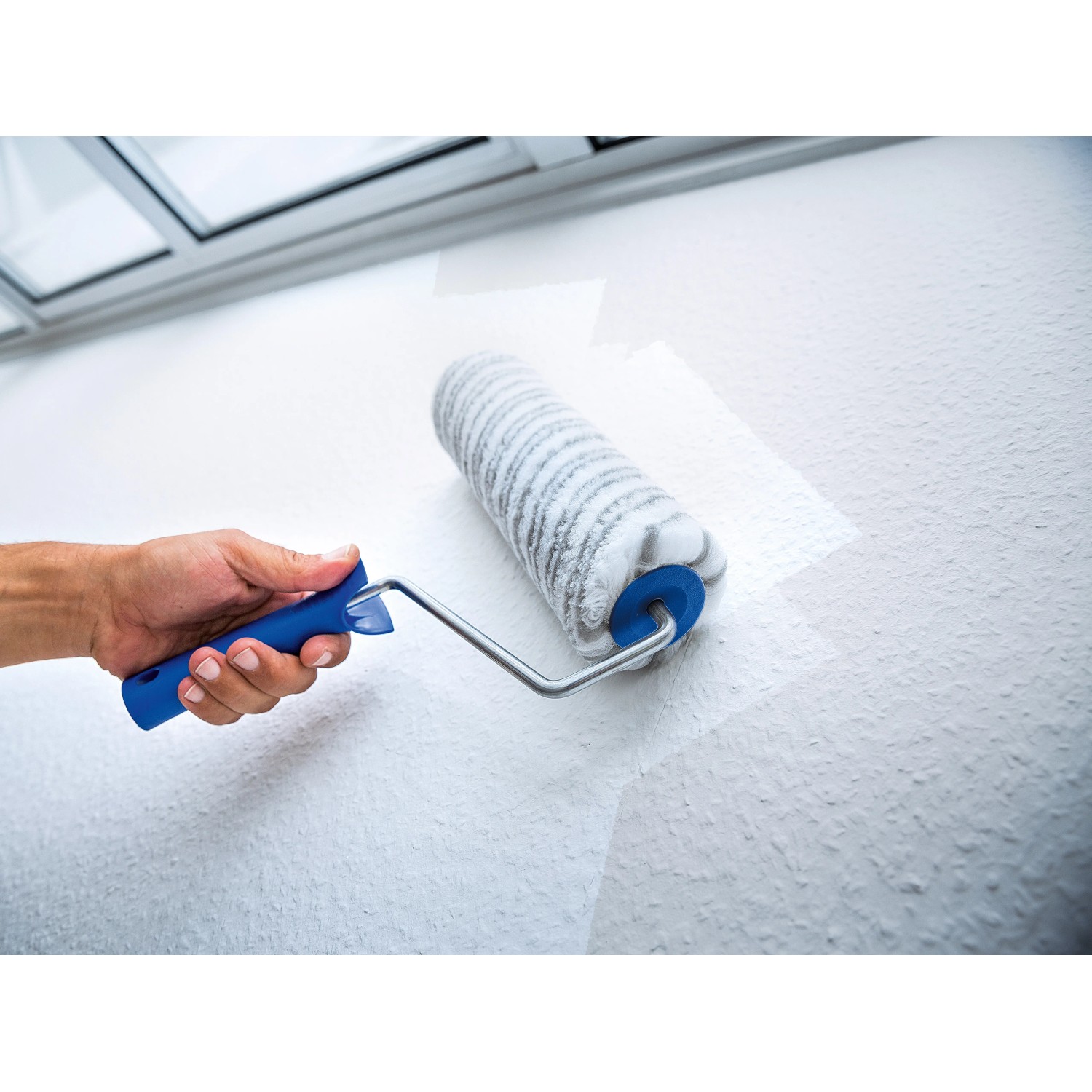 Rullo a dispersione LUX 18 cm mentre dipinge una parete. Rullo con manico blu e rivestimento grigio e bianco.