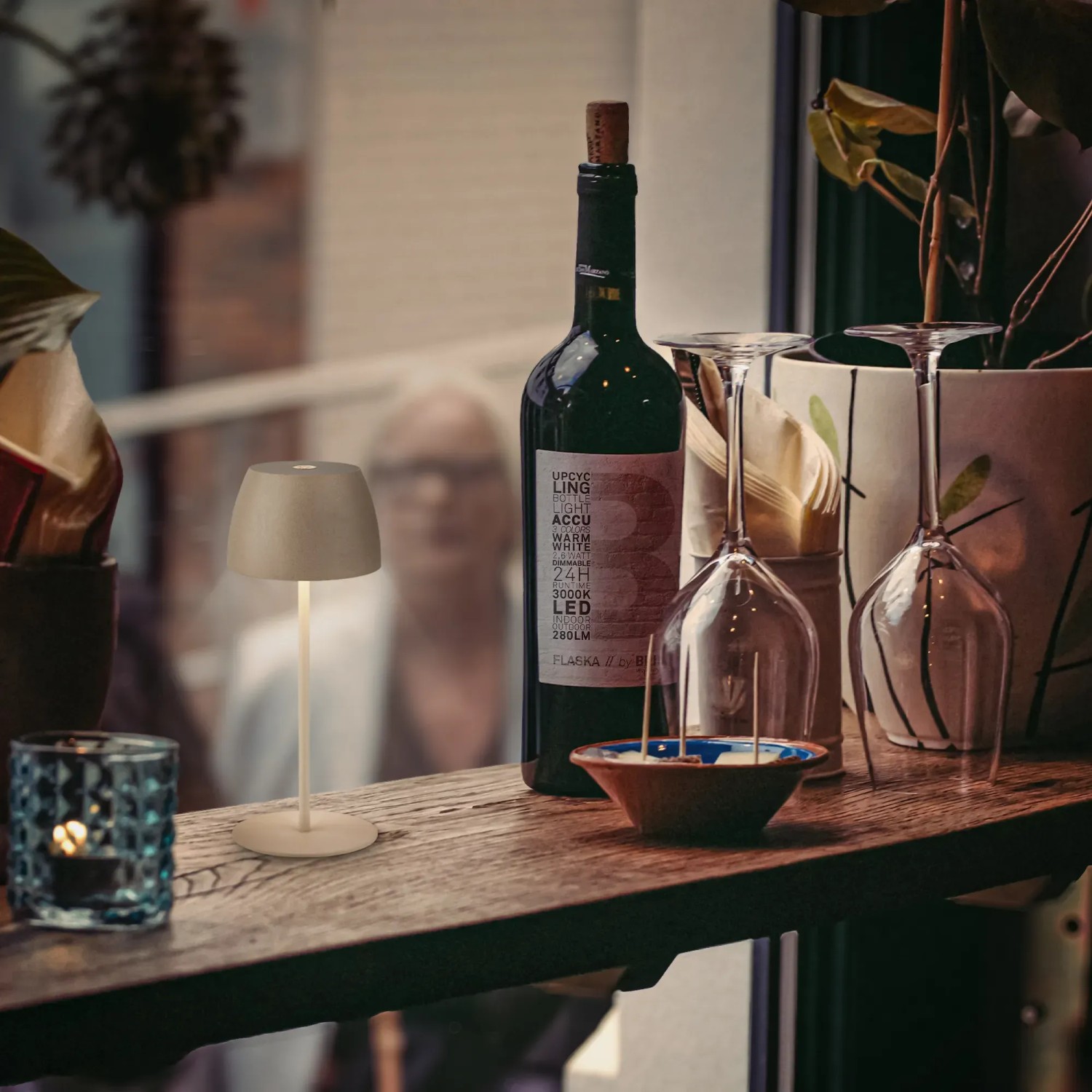 Lampada da tavolo a LED ricaricabile beige Brilo Serina Mini (20 cm) su tavolo con decorazioni.