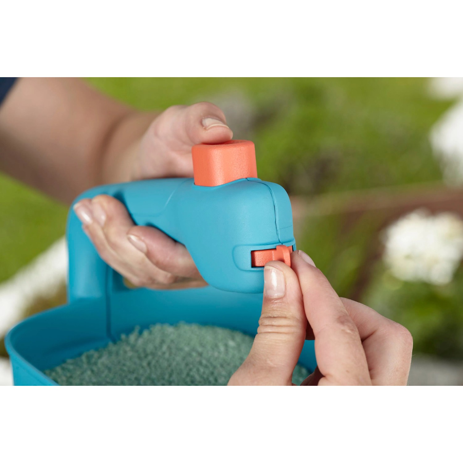 Primo piano: Mani azionano lo spargitore manuale Gardena M blu con semi, ideale per fertilizzanti e sale.
