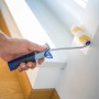 Mano che dipinge la parete con rullo a dispersione LUX 12 cm. Rullo con manico blu-grigio.