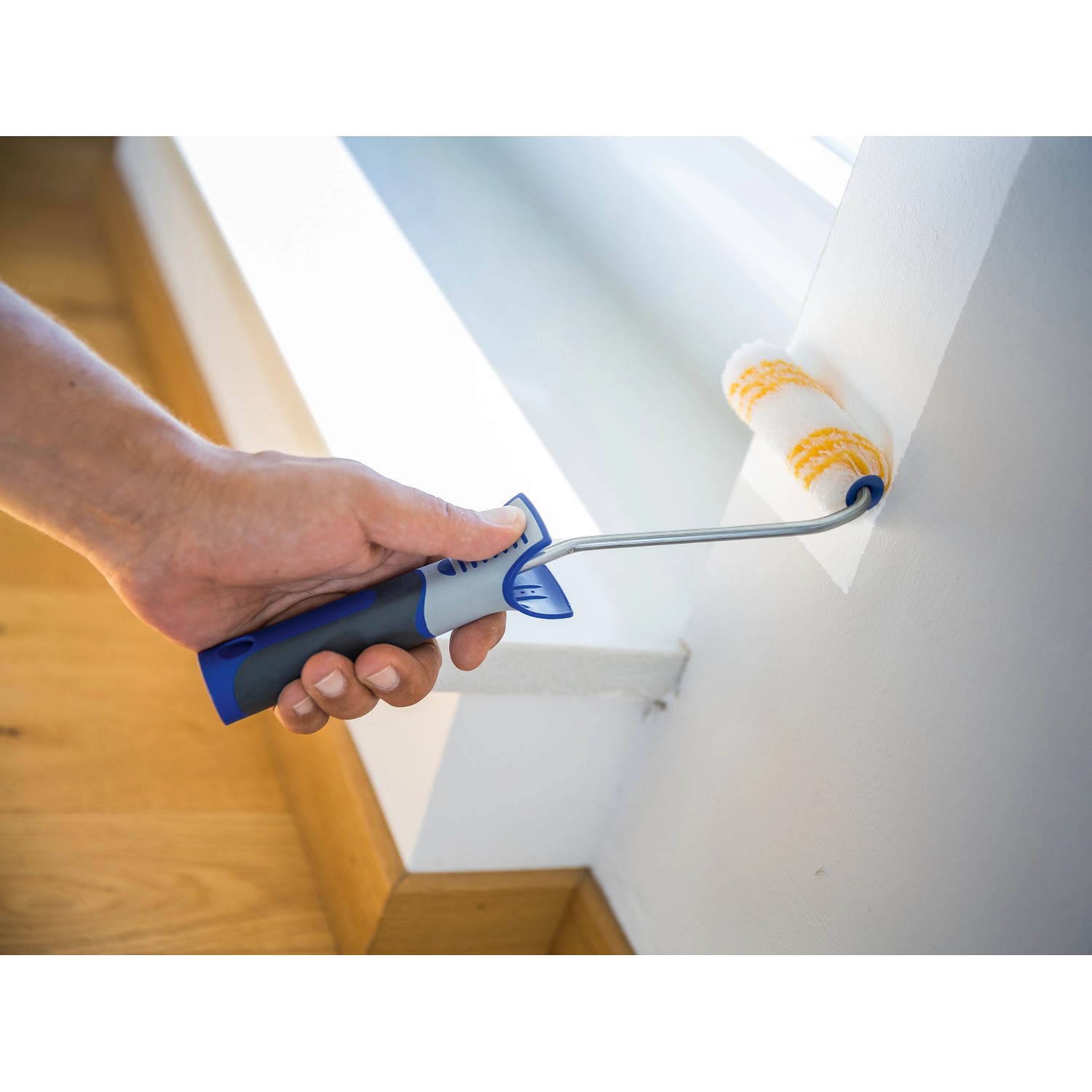 Mano che dipinge la parete con rullo a dispersione LUX 12 cm. Rullo con manico blu-grigio.