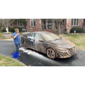 Uomo che pulisce un'auto molto sporca con un'idropulitrice davanti a una casa.