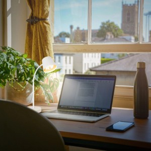 Lampada da scrivania a batteria Brilo Biwo bianca su scrivania con laptop e pianta.