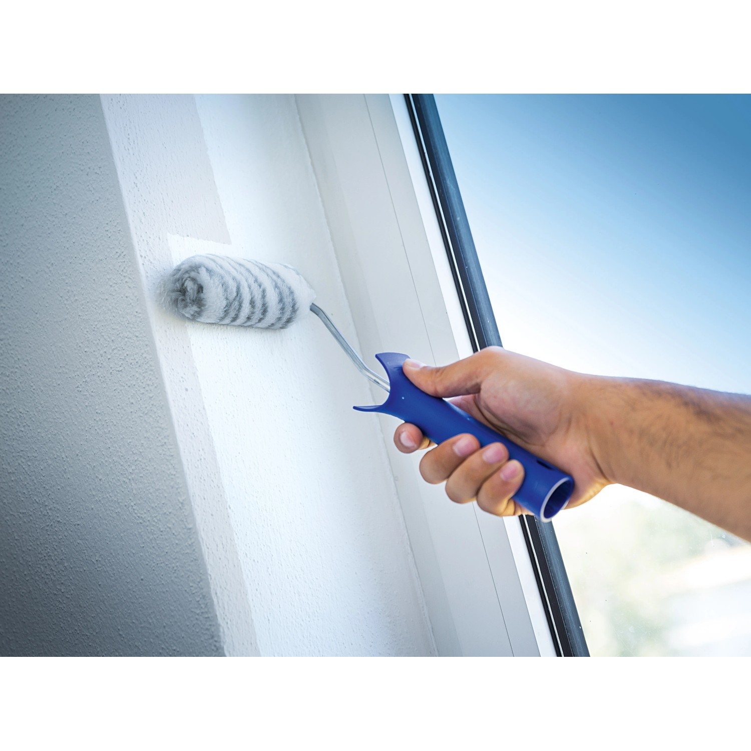 Mano che dipinge parete con rullo per dispersioni LUX 10 cm. Rullo con manico blu.