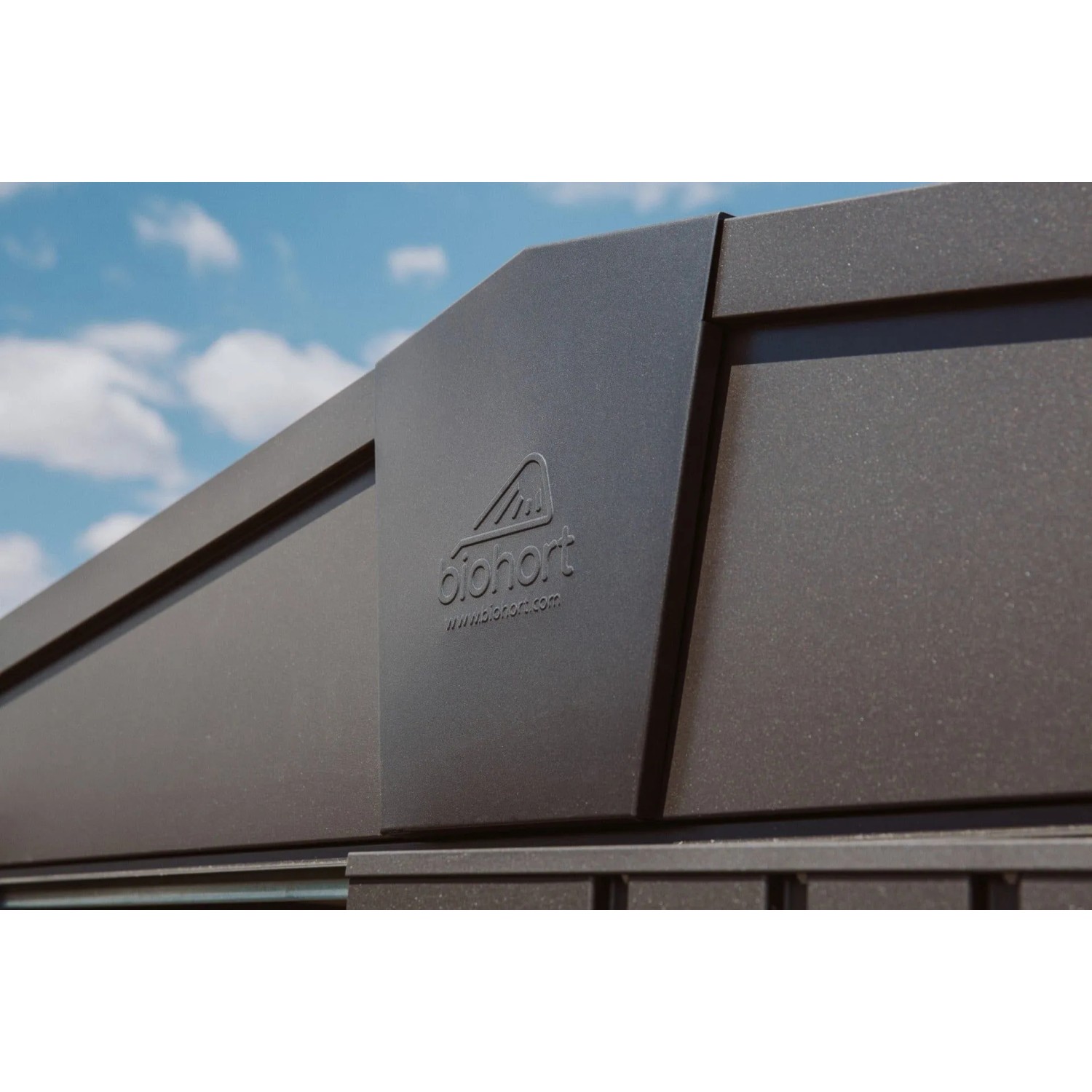 Casetta attrezzi Biohort Europa in metallo grigio scuro metallizzato. Parete robusta con logo Biohort sotto cielo blu.