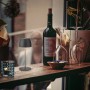 Lampada da tavolo a LED ricaricabile Brilo Serina Mini antracite su tavolo con bottiglia di vino e bicchieri.