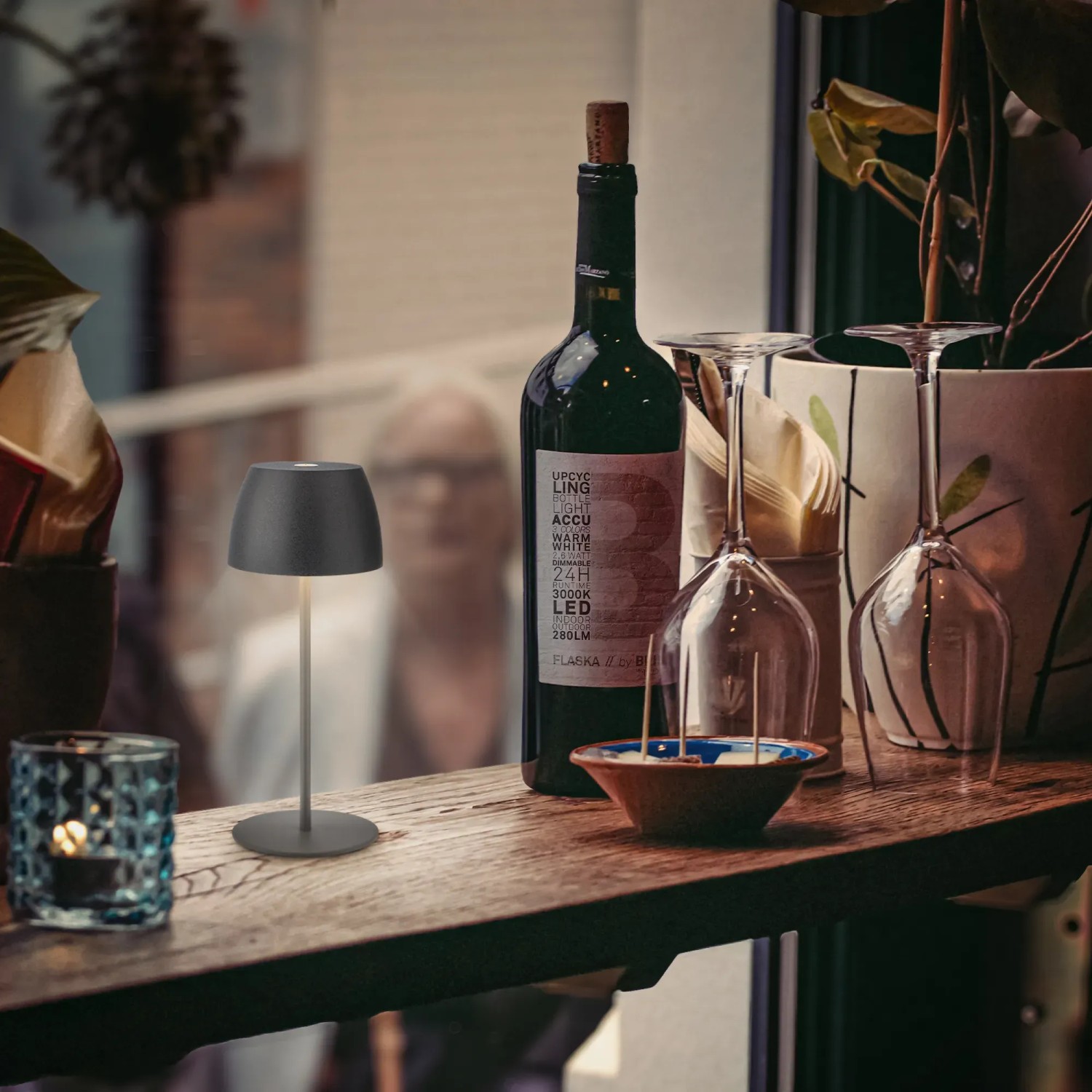 Lampada da tavolo a LED ricaricabile Brilo Serina Mini antracite su tavolo con bottiglia di vino e bicchieri.