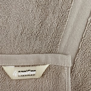Dettaglio di un tappeto da bagno beige con etichetta Saniflor. Texture morbida e finitura di alta qualità.
