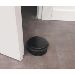 Fermaporta nero rotondo (Fermaporte) accanto a una porta su pavimento grigio.