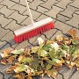 Scopa stradale LUX 40 cm con manico e setole rosse, accanto a un mucchio di foglie su pavimentazione.