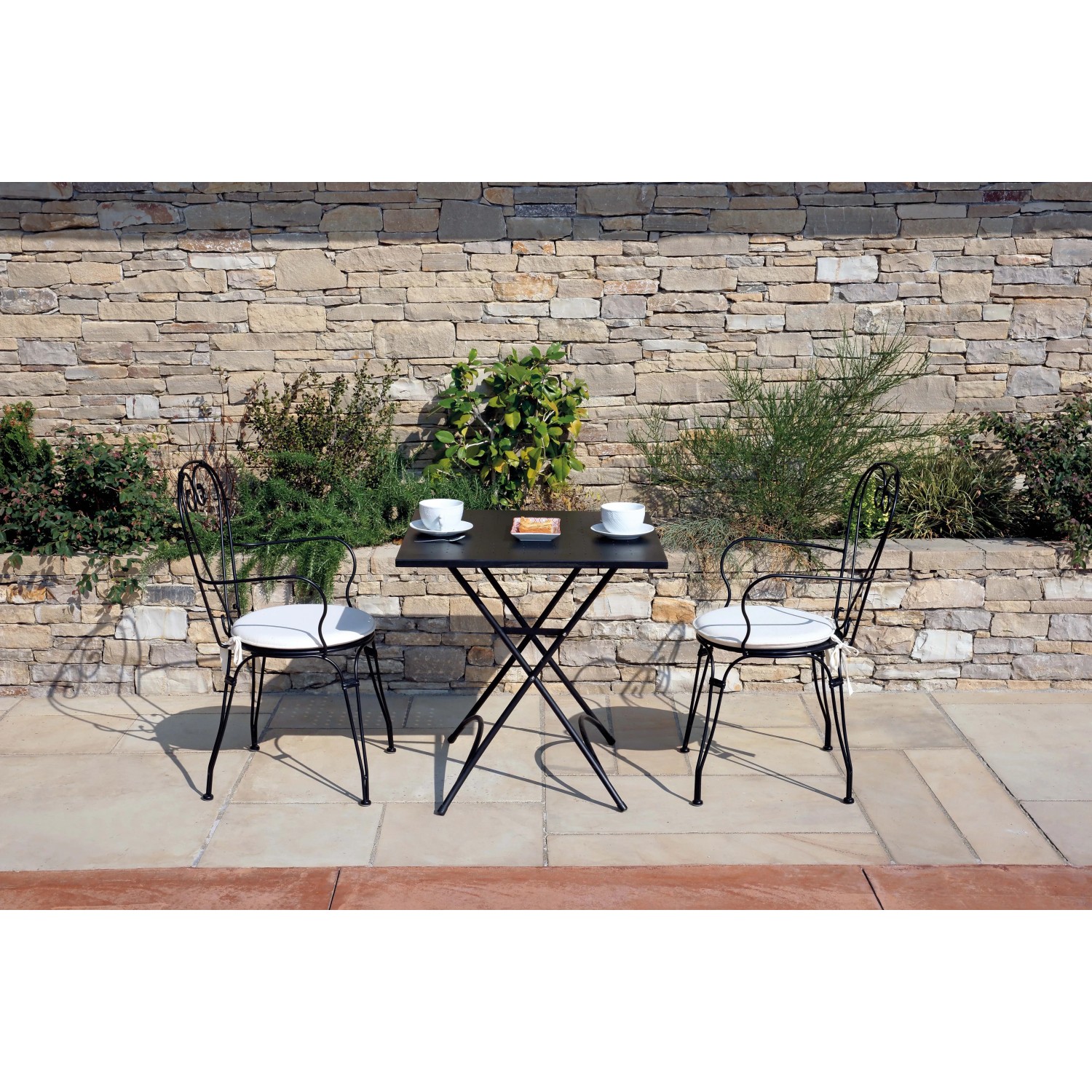 Tavolo da giardino nero con due sedie e cuscini bianchi davanti a muro di pietra.