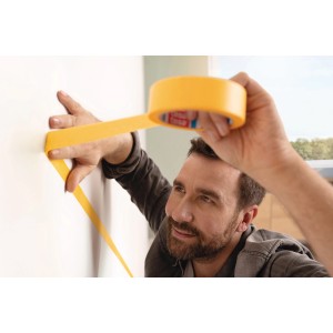 Uomo che applica nastro per pittori tesa® Malerband Perfect+ giallo su una parete bianca per bordi di pittura precisi.
