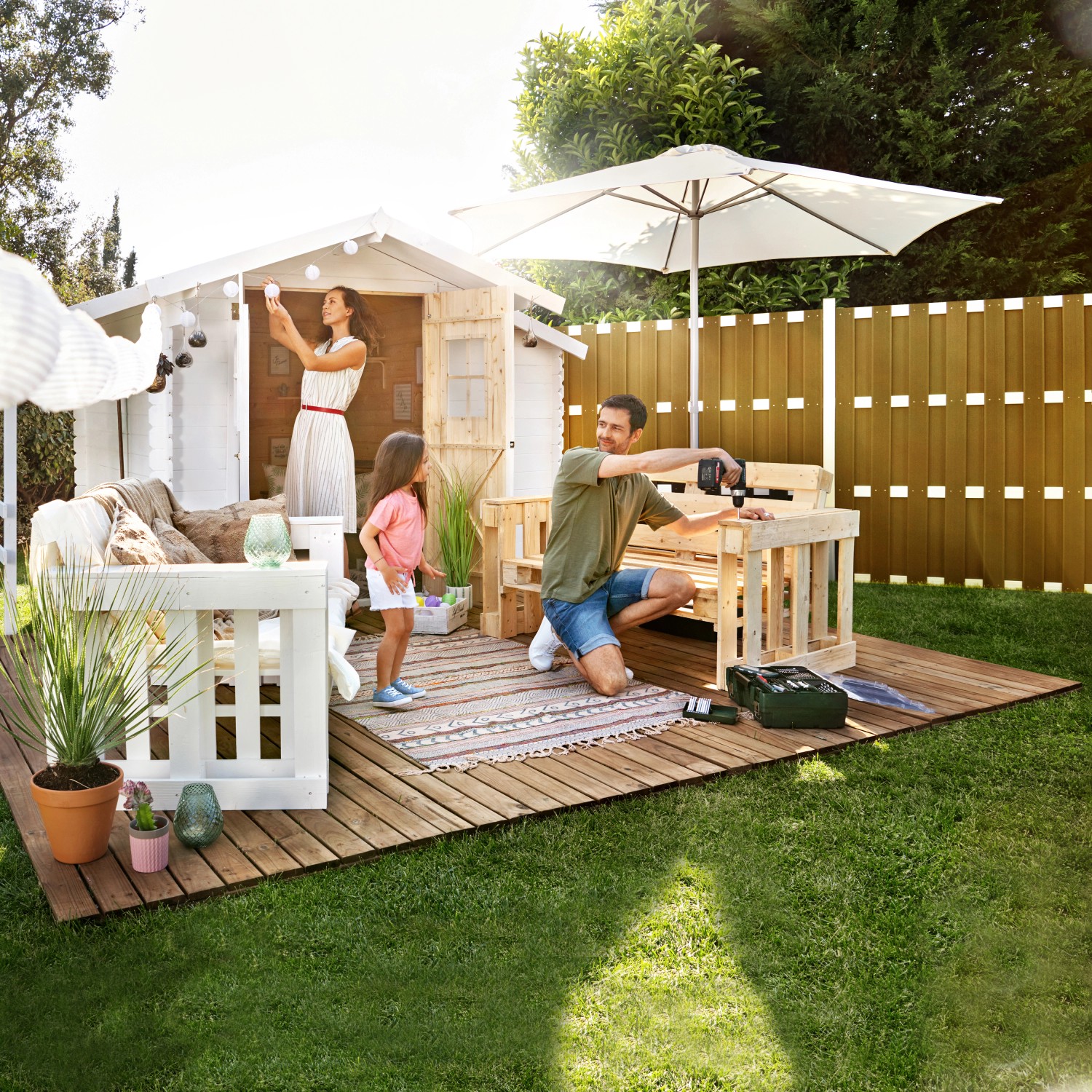 Famiglia che costruisce una recinzione privacy in WPC color sabbia in giardino con attrezzi e casetta da giardino.