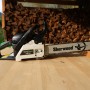 Motosega t.b.t. su tavolo di legno, catasta di legna sullo sfondo.