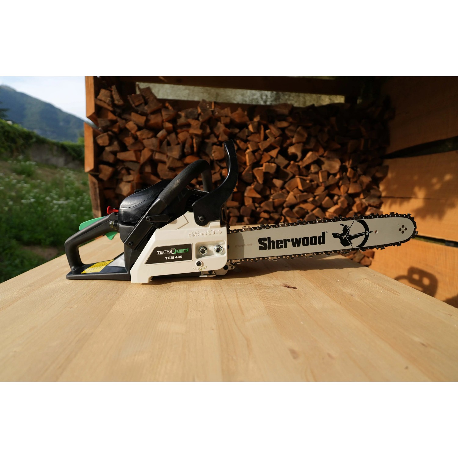 Motosega t.b.t. su tavolo di legno, catasta di legna sullo sfondo.