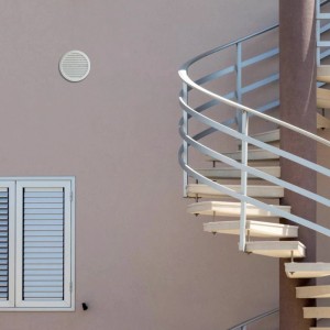 Griglia di ventilazione t.b.t. bianca e rotonda su una facciata accanto a una finestra e una scala a chiocciola.