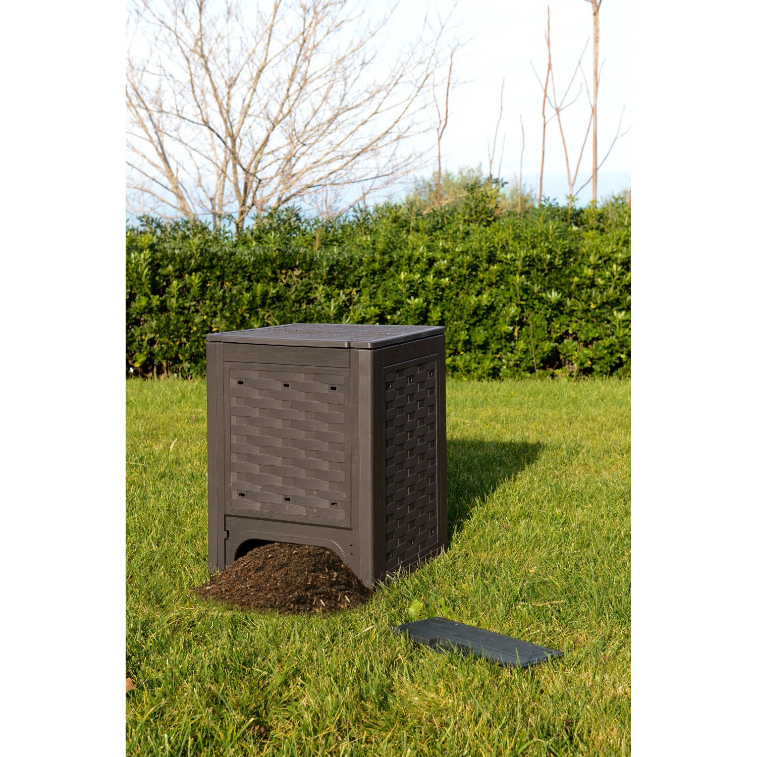 Compostiera t.b.t. marrone in giardino con terra di compostaggio davanti.