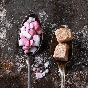 Due cucchiai: uno con zucchero colorato, uno con zucchero di canna. Quadri.