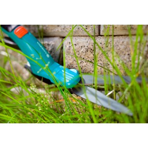 Cesoie per erba turchesi e grigie tra fili d'erba davanti a un muro di pietra.