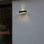 Lampada da parete solare a LED Lutec Fadi nera con sensore di movimento su muro grigio.