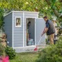 Casetta da giardino in plastica Keter, antracite, 1,8 m², con uomo che rastrella le foglie davanti alla casetta aperta.
