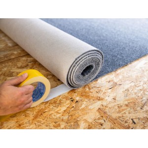 Mano applica nastro biadesivo LUX-TOOLS per pavimenti su pannello OSB accanto a tappeto srotolato.
