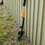 Erbacciaio ergonomico Fiskars, arancio-nero, appoggiato alla recinzione. Attrezzo da giardino per la rimozione delle erbacce.