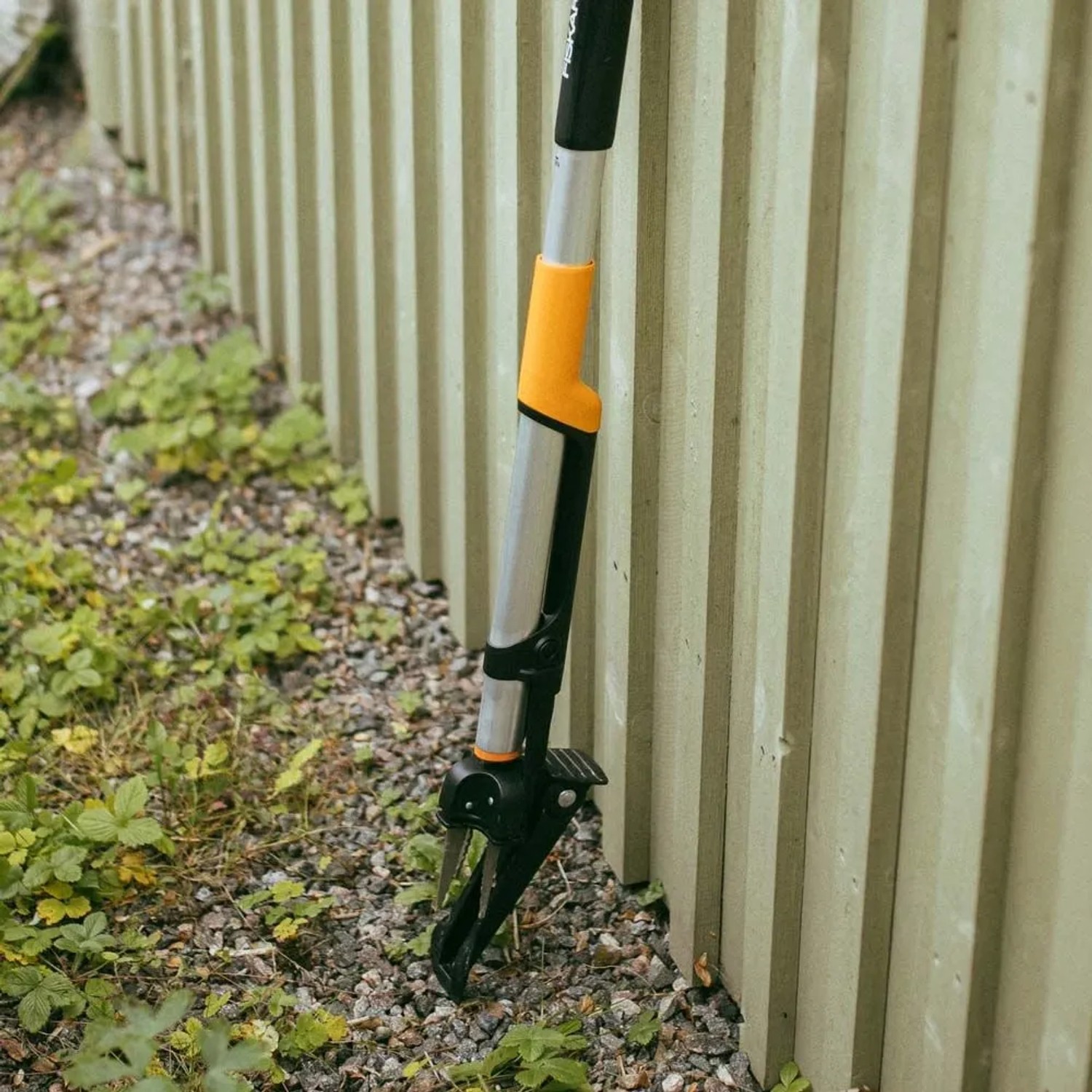 Erbacciaio ergonomico Fiskars, arancio-nero, appoggiato alla recinzione. Attrezzo da giardino per la rimozione delle erbacce.