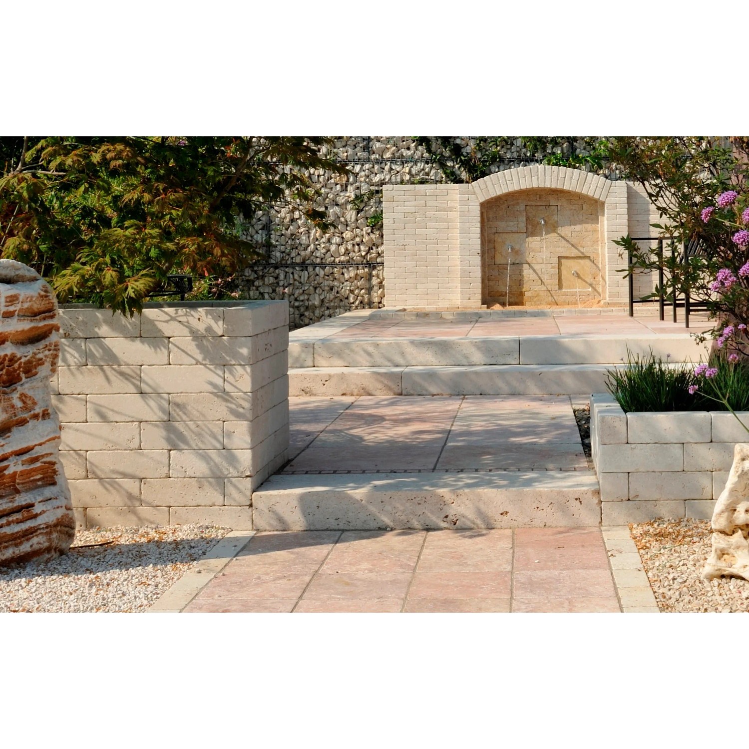 Giardino con elementi di muro e scale in pietra, bordura e fontana sullo sfondo.