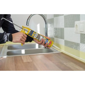 Persona sigilla lavello cucina con silicone bianco Soudal Cucina & Bagno. Pistola gialla su piano di lavoro in legno.
