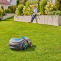 Robot tagliaerba grigio su prato, uomo con smartphone sullo sfondo.