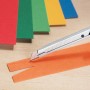 Cutter LUX 9 mm in metallo taglia carta arancione, pila di carta colorata sullo sfondo.