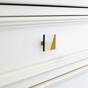 Pomello per mobili quadrato con motivo geometrico in bianco, beige e oro su cassetto bianco.
